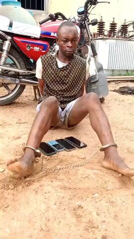 A man is sitting on the ground, handcuffed at his wrists and ankles. He is wearing a patterned vest over a white t-shirt and light-colored shorts. In front of him are four mobile phones laid out on the dirt. Behind him is a red motorcycle and a building. The ground is sandy and dry. The man appears to be looking down.