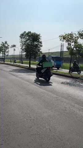 The video shows a person riding a motorcycle on a paved road. The motorcycle has a green box strapped to the back. There are trees and streetlights along the side of the road. A blue truck is visible in the background. The sky is clear and the sun is shining.
