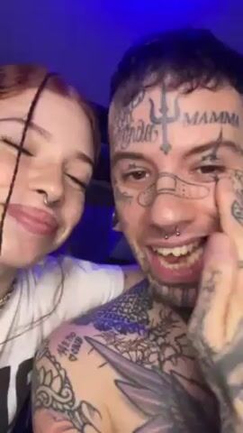 A close-up shot shows a man and woman posed intimately close together. The man is fair-skinned and heavily tattooed on his face, neck, chest, and arms. The tattoos include words like “MAMMA,” various symbols, and decorative designs. He has gauges in his ears and several piercings in his nose. He is shirtless. The woman has fair skin, light red hair in long braids, and a nose ring. She wears a white t-shirt. Both look into the camera. The background is a blurred out blue color. The man is holding his hand up to his face at times, obscuring parts of his cheek and eye.