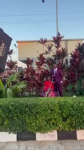 The scene is an outdoor photoshoot. A couple is posing among plants; the woman is wearing a red dress, and the man is wearing a purple suit. A black softbox on a stand, labeled "Godox", is positioned to their left. A photographer, wearing a black t-shirt and cap, is taking pictures. Another light modifier, an umbrella, is set up nearby on a stand. In the background, there is a building, a tall pine tree, and a security camera mounted on a pole. The ground is paved with light-colored brick. The sky is light blue. The photographer gives directions to the couple.