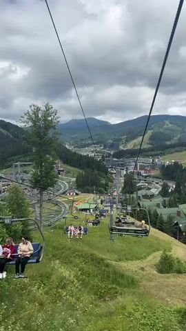 The video shows a view from a ski lift in a mountainous area with a village nestled in the valley below. The ski lift is carrying passengers up a grassy slope, and several people are visible sitting in the chairs. The village below consists of many buildings, some of which appear to be ski resorts or hotels. There are also ski slopes and roller coaster tracks visible in the distance. The sky is cloudy, and the weather appears to be overcast.