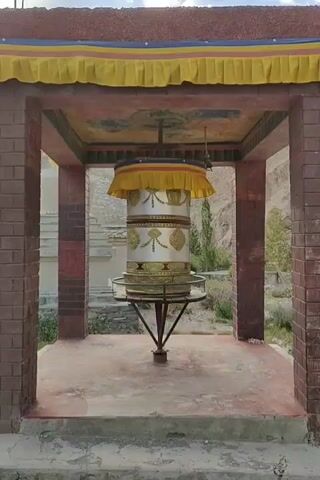 A prayer wheel spins within an open structure with brick pillars. The prayer wheel is cylindrical and ornate, with gold-colored embellishments against a white background. It is topped with a yellow and red fabric canopy and hangs from a central support. The structure has a painted ceiling and a worn, reddish-brown floor. In the background, a white building with decorative elements is visible, along with dry vegetation and a mountainous landscape. The sky is partly cloudy. The video captures the gentle rotation of the prayer wheel.