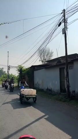 The video shows a street scene with a person pulling a cart. The cart contains several large, white, rectangular containers, possibly water jugs. The person is walking behind the cart on a paved road. In the background, there are several motorcycles carrying people, a building with a light-colored wall, and a utility pole with numerous wires. The sky is clear and the day appears to be sunny. The environment appears to be a rural or semi-urban area.
