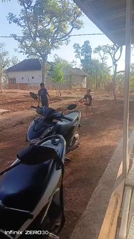 In a rural outdoor setting in Sokaramme Timur, Indonesia, two young boys are engaged in a playful activity. The scene is bathed in the warm light of what appears to be late afternoon or early morning. The ground is a mixture of dry earth and scattered leaves, suggesting a somewhat arid environment.

Dominating the foreground is a modern black scooter, its sleek design contrasting with the rustic surroundings. Behind the scooter, the two boys are actively interacting. One boy, dressed in a dark t-shirt and shorts, is seen in motion, possibly running or jumping. The other boy, wearing a lighter-colored t-shirt and shorts, appears to be holding something and is also in an active pose. Their movements suggest a game or a chase.

In the background, traditional Indonesian houses with light-colored walls and dark roofs are visible, nestled amongst lush green trees. The trees are tall and leafy, providing shade and a sense of established nature. The overall atmosphere seems peaceful and typical of a village in Southeast Asia.

The sky is a clear, pale blue, indicating fair weather. There are no overt signs of distress or conflict among the children; their engagement appears to be one of carefree play. The perspective is from under a covered structure, possibly a porch or veranda, with bamboo elements visible along the edge of the frame.

The timestamp "00:00" is visible in the top left corner, indicating the start of a recording. In the bottom left corner, the text "Infinix ZERO30" is present, which is likely the model of the device used to capture the video. This detail suggests the video is a mobile phone recording.