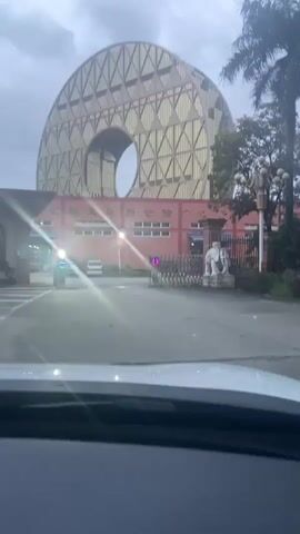 The video shows a large, golden, donut-shaped building with a hole in the center, seen from a car's windshield. The building has a reddish-brown facade with windows. There are palm trees and other foliage to the right of the building. In front of the building is a closed metal gate, flanked by two white elephant statues. A traffic light is visible near the gate. The sky is overcast and appears to be evening or early morning. There are some vehicles parked on the left side of the road. The footage appears to be shaky, suggesting it was recorded by someone in a moving vehicle.