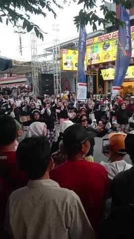A crowd gathers outdoors for what appears to be a performance or event. In the foreground, the backs of several individuals are visible, facing away from the camera. They are dressed in casual clothing, including a red shirt, a patterned shirt, and a red baseball cap. Beyond them, a large group of people are assembled, many wearing headscarves, suggesting a religious or cultural context. Some individuals in the crowd are holding up their phones, likely to record the event. In the background, a stage with scaffolding and speakers is set up, indicating a performance area. Banners and signs are visible, some featuring advertisements. The overall impression is of a lively public gathering or festival.