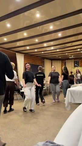 A group of young people, mostly women and a few men, are dancing in a lively circle inside what appears to be a dining hall or common room. The room has a wooden-beamed ceiling with recessed lighting, light-colored walls with white curtains, and a television mounted on one wall. Tables with chairs are visible in the background, some covered with white tablecloths.

The individuals are dressed in casual attire, including t-shirts, jeans, and comfortable pants. They are holding hands and moving rhythmically to the music, smiling and laughing, indicating a cheerful and social atmosphere. One person has the name "ИВАНОВА" (IVANOVA) printed on the back of their black t-shirt. The setting suggests a social gathering or event in Yus'ki, Russia.
