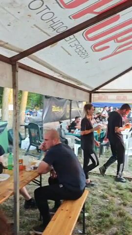The video captures an outdoor gathering, likely a motorcycle rally or event, with people seated at tables under a large tent. The tent has a white canopy with red lettering that appears to be a brand logo, possibly of a beverage company. In the foreground, a man with a shaved head sits on a wooden bench. To his right, a couple walks past, the woman holding a drink. Further in the background, numerous motorcycles are parked. People are seated at various tables, some eating or drinking. The ground is covered with grass and some scattered dirt. The overall atmosphere suggests a casual, social event.