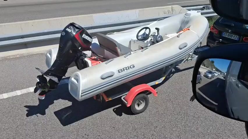 The video shows a gray inflatable boat on a red trailer, with an Evinrude 35 motor attached, parked on the shoulder of a highway. Traffic is visible in both directions, with several cars passing by. A concrete barrier is next to the boat. The vehicle recording the video is parked next to the boat.