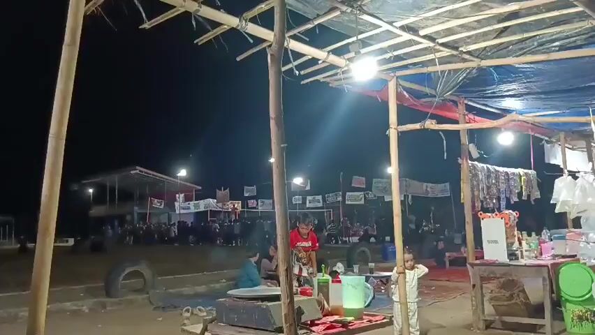 The video shows an outdoor night scene, likely a market or fair. In the foreground, there's a makeshift food stall with a table covered in various items, including bottles of condiments, a pitcher, and possibly some food being prepared on a hot plate. A young boy in a red shirt and two younger children are near the table. Behind the stall, there's a gathering of people sitting and standing, some under a covered structure with lights. Further in the background, more stalls or structures are visible, adorned with banners or flags. The ground appears to be dirt or sand. The overall atmosphere seems casual and lively, with artificial lighting illuminating the area.