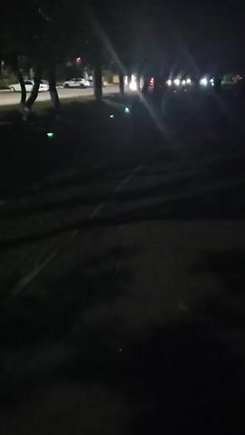 The video is a low-light recording taken at night, likely from a moving perspective as the scene is blurred. It appears to be an outdoor setting with trees lining what looks like a path or road. Several small green lights are visible along the edge of the path, and further in the distance, there are blurry lights that suggest moving vehicles or streetlights. The overall atmosphere is dark and somewhat obscured by motion blur and low light conditions.
