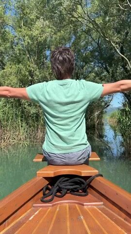 A person is seated in a wooden boat, facing away from the camera, on a narrow waterway surrounded by green reeds and trees under a clear sky. The person is wearing a light green t-shirt and grey shorts. Their arms are initially extended horizontally. The person then turns their head to the left, looks towards the camera, makes a kissing face, and raises their left hand with the middle finger extended. They then return to the initial pose with arms outstretched, facing away from the camera. A black rope is coiled in the front of the boat.
