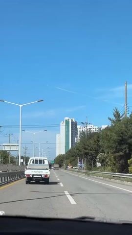 This video captures a view from inside a vehicle traveling on a multi-lane road in Cheongju-si, South Korea. The time of day appears to be late morning or early afternoon, given the bright sunlight and clear blue sky with a few wispy clouds and a contrail from an airplane. The weather seems pleasant and sunny.

The primary subjects are a white Hyundai truck in the foreground and several tall residential buildings in the background. The truck is moving ahead of the camera vehicle in the right lane. The buildings are modern high-rise apartment blocks, one of which prominently features a green vertical stripe and the word "SK" in white lettering. Other smaller buildings and structures are also visible, including utility poles with many wires and what appears to be a cell tower disguised as a tree.

The setting is a paved road with clear lane markings, including a yellow line separating lanes and white dashed lines. Guardrails line the side of the road, and there is a grassy area with trees and bushes on the right. Streetlights are visible along the road.

The activity is simply driving along the road. There are a few other vehicles on the road, including cars and trucks, but the focus is on the road ahead and the surrounding environment.

Notable details include the "HYUNDAI" logo on the back of the truck, the "SK" branding on the building, and a Korean road sign with blue and white text on the right side of the road. The overall mood is calm and ordinary, depicting a typical daytime commute or journey.