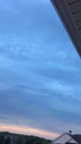 A low-angle shot shows a blue sky with scattered clouds. Two airplanes are visible in the distance. The first airplane, which is smaller, is slightly to the left of the frame. The second airplane, which is larger and appears to be a commercial jet, is closer to the center of the frame, with its navigation lights visible. The video briefly zooms in on the larger airplane as it flies through the clouds. The shot ends with a view of the airplane from a slightly different angle as it continues to fly.