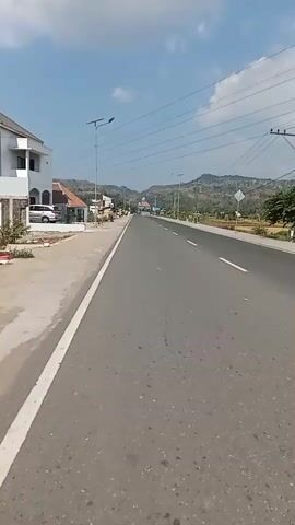 The video captures a view from a moving vehicle traveling on a paved road. On the left side of the road, there is a sidewalk and several buildings, including a white house and smaller structures. Beyond the sidewalk, a few vehicles are visible, including a silver car. The road itself is a two-lane highway with a white dividing line and a white edge line. Telephone poles with power lines run along the side of the road. In the distance, the road continues towards hills under a clear blue sky with some scattered clouds. There are streetlights along the road. Some areas off the road appear to be fields or undeveloped land. The overall impression is a rural or suburban roadside scene on a sunny day.