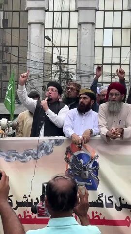A man in a white shirt and vest, wearing a black cap, speaks into a microphone while gesturing with his left hand. He is standing on a raised platform behind a banner that features a chained lock superimposed on an orange and blue emblem. Several other men are also on the platform, some wearing traditional religious attire and caps, and some are holding up a poster with images. The man speaking is the central focus, with many other individuals in the background and foreground, some holding cameras. The environment appears to be an outdoor urban setting with buildings and electrical wires visible. The banner is partially visible at the bottom of the frame, with text in a non-English script.