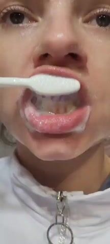 A close-up shot shows a person's mouth with a toothbrush covered in white toothpaste and foam. The person is in the process of brushing their teeth. The toothbrush is white with a purple tip. The person's lips are pink and slightly parted. The background is blurred and appears to be a white wall or surface.