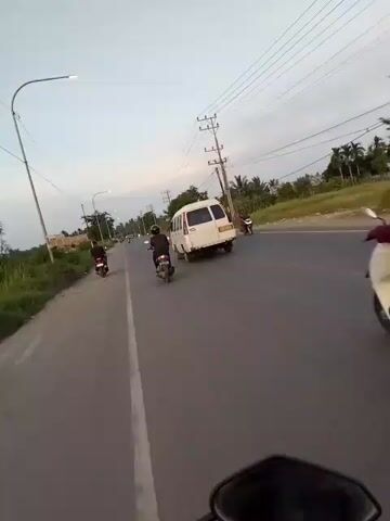 A point-of-view video captures a motorcycle riding on a two-lane road. The road appears to be paved and has a white line marking the lanes. The surrounding environment is rural, with fields of grass and some trees visible on the left side of the road. On the right side, there are more trees, some buildings, and utility poles with power lines stretching across the sky. Several other vehicles are on the road, including three motorcycles and a white van. The motorcycles are all in motion, with one rider in front of the camera, one slightly ahead and to the left, and another further ahead and to the right. The white van is also in front of the camera, traveling in the same direction. The sky is overcast, indicating it might be dawn or dusk. The video is shot from a low angle, suggesting the camera is mounted on the motorcycle's handlebars or dashboard.