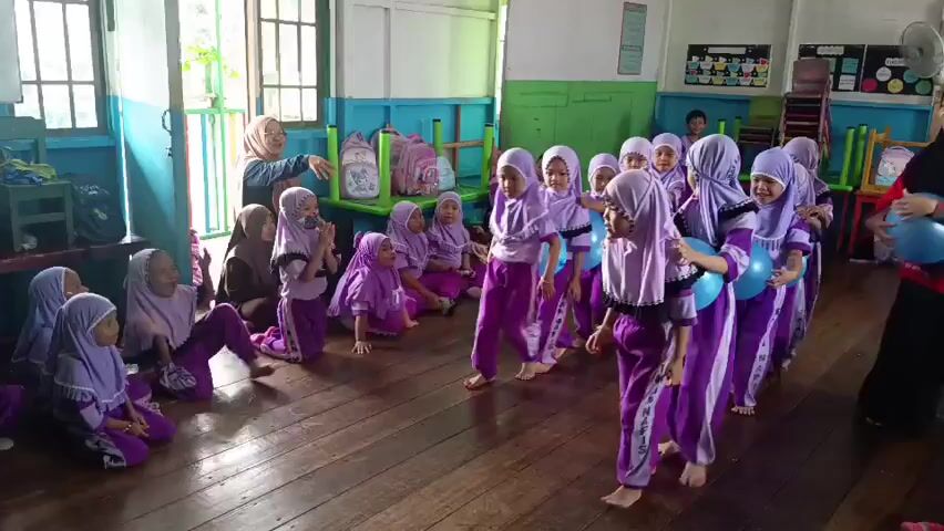 A group of young girls in purple hijabs and matching outfits are playing a game with blue balloons in a classroom. Some girls are standing in a line, holding balloons between their knees, while others are sitting on the floor. A woman in a red shirt and black pants is standing to the right, seemingly facilitating the game and holding a balloon. Another woman in a hijab and brown dress is standing in the background to the left, also appearing to supervise. The room has wooden floors, blue and green painted walls, windows, and some colorful bags and chairs visible. The overall atmosphere appears to be one of fun and educational activity for the children.