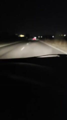 A first-person perspective video shows driving on a dark, empty road at night. The road has a white line dividing the lanes. Headlights from the car illuminate the road ahead. In the distance, several cars can be seen with their taillights illuminated. One car directly ahead is clearly visible, its red taillights and license plate illuminated. The surrounding environment is dark with only faint lights visible on the horizon, suggesting a rural or undeveloped area.