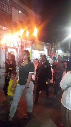 Here is a description of the video:

The video appears to have been taken during a nighttime celebration or parade. In the initial shot, there are flaming torches on what looks like a mobile unit. A crowd of people walks along a street in front of buildings. 

Subsequent shots show people walking, and a group of men in matching patterned shirts with musical instruments, including drums, heading in the same direction. There are motorbikes parked on the street. It is difficult to distinguish specific details due to the lighting, but the video seems to document a lively, festive event.