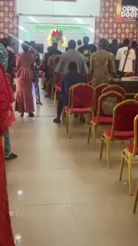 The video shows a gathering of people inside what appears to be a church or auditorium. People are milling about, standing, and sitting in rows of red chairs. Some are dressed in formal or traditional attire. A stage area is visible in the background, with a sign that says "OPEN DOOR." A screen above the stage displays text, potentially lyrics or announcements. The overall impression is that this is either a pre-service or post-service gathering, or perhaps an intermission.