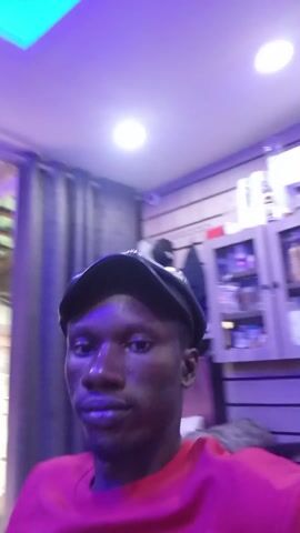 A man with dark skin, wearing a red t-shirt and a black hat, is looking directly at the camera. He is indoors, under bright, slightly purplish lighting. In the background, there is a shelf filled with objects, a window or glass door, and what appears to be a partially visible mannequin. The video is slightly shaky.