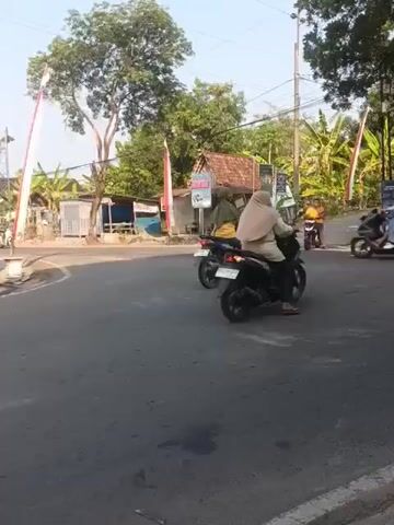 A short video captures a street scene with multiple motorcycles on an asphalt road. In the foreground, a blue motorcycle is partially visible. Behind it, a black motorcycle with a rider wearing a light-colored headscarf is moving. Further back, a dark blue motorcycle with a rider in a green jacket is also in motion. Another black motorcycle with a rider is visible in the distance. The background shows a small building with a red roof, trees, and tropical plants, including banana trees. Red and white flags are mounted on poles on either side of the road. Power lines are visible overhead. The lighting suggests it is daytime, possibly late afternoon, with shadows cast on the road.