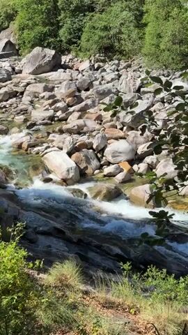 The video shows a clear, turquoise river flowing over a bed of smooth, grey, and tan boulders. The water is fast-moving, creating white foam as it cascades over the rocks. The river is surrounded by lush green trees and foliage on either side, with larger boulders and rock formations visible further up the embankment. The sunlight filters through the trees, casting dappled light on the water and rocks. There are no people visible in the immediate scene. The overall impression is a natural, serene landscape.