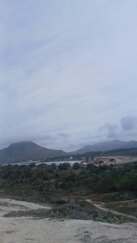 The video presents a scenic overlook of a coastal landscape under a cloudy sky. Mountains form the backdrop, with one large peak partially obscured by clouds on the left side of the frame. In the midground, a beach is visible along the coastline, where at least one person appears to be kitesurfing. The foreground is characterized by scrubby vegetation including trees and cacti, which lead to a sandy terrain on which two figures can be seen walking along a path. The overall tone of the video is subdued, due to the overcast lighting, but the kitesurfer adds a splash of color and activity to the scene.
