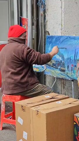 An artist sits on a red plastic stool on a sidewalk, painting. He wears a red cap, a brown jacket, and tan pants. The artist is working on a painting of a steam train in a rainy environment, complete with figures holding umbrellas. His palette is in his left hand, and a paintbrush in his right, as he adds details to the painting. In front of the artist are two brown cardboard boxes. Behind him is a wall, a window, and other painted canvases. It sounds like someone is calling the artist's name.
