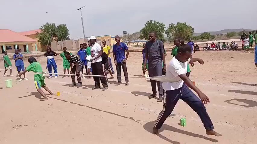 The video shows a game in progress on a sandy field with several people looking on. The people appear to be a mixture of children and adults. The game appears to be a limbo-type activity, where participants must go under a pole without touching it. One person is shown in the foreground attempting to get under the pole, while another is in the background. Several people are standing around the pole, possibly supervising or watching. The background includes buildings, trees, and a light pole. The sky is overcast.
