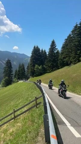 The media file features a scenic motorcycle ride through the Italian Alps, specifically in the area of St. Magdalena - Santa Maddalena.

The primary subjects are a group of motorcycles and their riders, navigating a winding asphalt road that cuts through a lush, green mountain landscape. The motorcycles appear to be sport or adventure bikes, equipped with riders in full protective gear, including helmets.

The scene is set against a backdrop of majestic mountains covered in dense pine forests and rolling green hills, characteristic of the Dolomites. A charming village with scattered houses is visible nestled in a valley below, adding to the picturesque setting. The perspective shifts, offering panoramic views of the dramatic mountain peaks in the distance, some with jagged, rocky summits. Wooden fences line the roadside in some shots, further emphasizing the rural and natural environment.

The activity taking place is a recreational motorcycle journey, likely a tour through scenic routes. The time of day appears to be midday or early afternoon, given the bright sunlight and clear blue sky punctuated by fluffy white clouds. The weather is fair and pleasant, ideal for outdoor activities like motorcycling.

Notable details include the vibrant green of the grass and trees, contrasting with the dark green of the forests and the grey of the distant mountain ranges. The clear, bright lighting suggests a sunny day, contributing to the overall cheerful and adventurous mood. The road itself is well-maintained, with clear lane markings and a metal guardrail.

There is no visible text in the media file. The overall impression is one of freedom, adventure, and the breathtaking beauty of the Italian countryside.