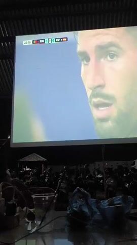 This media file shows a projection screen displaying a soccer match, likely the FIFA World Cup, as indicated by the text "FIFA WORLD CUP QATAR 2022". The score is Portugal 0, Spain 0 at 22:10. The screen shows a close-up of a soccer player's face, followed by a wide shot of the soccer field with players on the field and a large audience in the stadium.

In the foreground, there are several objects on a table, including a glass of liquid with a straw, a small bottle, and a crumpled plastic bag. The scene appears to be set outdoors at night, with a dark ceiling overhead and dim lighting. The presence of a crowd gathered to watch the match suggests a social event, possibly in a public space like a bar or restaurant in Miruektaman, Indonesia, where people are watching the game on a large projected screen. The overall mood seems relaxed and engaged, with the audience focused on the game.