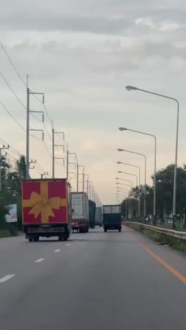 A medium shot shows a highway with a line of trucks driving on it. The sky is overcast, and the road is gray. The trucks are all facing away from the camera, and they are all driving in the same direction. The first truck, on the left, is red with a yellow ribbon on it, resembling a gift box. The other trucks are various colors and appear to be shipping containers or box trucks. Streetlights line the right side of the road. Utility poles with power lines are visible on the left side of the road.