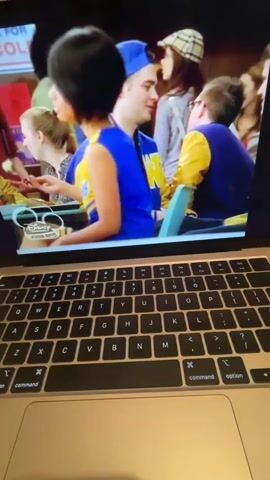 A young woman with short dark hair, wearing a yellow and blue cheerleader outfit with "HOLLANDS" written on it, sits in the foreground. She holds a fork with a piece of food up to her mouth. Behind her, a young man wearing glasses and a yellow and blue jacket looks in her direction. In the background, a banner reading "HOLLAND VOLLEYBALL" is visible. The scene appears to be a sporting event or school gathering. A laptop keyboard is visible in the bottom half of the frame.