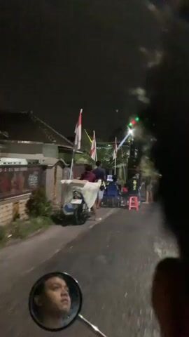 The video shows a nighttime street scene with several people gathered around a large sound system and stage setup. There are Indonesian flags displayed. The camera appears to be mounted on a moving vehicle, likely a motorcycle, as a reflection of the rider is visible in a mirror. People are seen standing, sitting, and interacting. Some individuals are near a laptop and sound equipment, suggesting they are operating the audio system. The stage is illuminated with colorful lights, and multiple speakers are stacked. The environment appears to be a residential or commercial area with buildings visible in the background. The overall atmosphere suggests an outdoor event or gathering.
