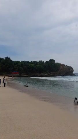 The video shows a beach on a cloudy day. The sand is light-colored, and the water is clear with small waves. There are rocky cliffs covered in trees on either side of the beach. Initially, a lone woman sits in the water near the shore. Later, a group of four people, two adults and two children, are seen wading in the water. One of the adults walks deeper into the water away from the group. In the distance, a large rock formation is visible in the sea.