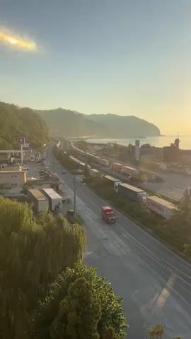 The video provides an aerial view of a coastal highway and train track during a sunset. A red truck is driving on the highway. A train is parked on the train track. There are buildings and trees on the left side of the highway. The ocean and hills are visible in the background. The sun is setting, casting a warm glow over the scene. A faint rainbow-like optical phenomenon is visible in the upper left corner of the sky.