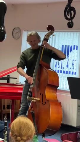 A man plays a double bass in a room with children. He is standing and holding the neck of the instrument with his left hand and bowing it with his right. He is wearing a dark green sweater and blue jeans. In the foreground, the backs of several children's heads are visible. One child has blonde hair in a braid. In the background, a red piano or keyboard is visible to the left. A clock is on the wall. A window with blinds is behind the man. Music notes are hanging from the ceiling. The room appears to be a music classroom or studio.