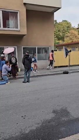 A group of people are gathered on a sidewalk next to an apartment building. One person is kneeling on the ground and taking a photo with a camera. Another person is standing and holding a pink umbrella. Two other people are standing near a building entrance. A person is standing on a skateboard on the sidewalk. A camera light is set up on a stand in the background. The scene appears to be a photo shoot.