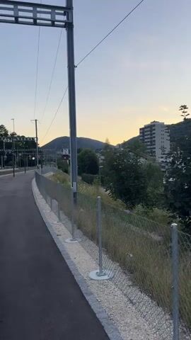 The video pans across a somewhat rural-urban fringe scene at dusk in Moutier, Switzerland. The foreground features a paved path bordering a chain-link fence. Beyond the fence, overgrown grass and weeds grow along a slope, with tall trees and bushes interspersed with buildings.

In the midground, a modern, multi-story apartment building with balconies and many windows stands out against a wooded hillside. To the left of this building, a lower, older concrete structure is visible. Further to the right, another apartment building with a lighter facade is situated. A red car is parked on a paved area near some trees. A lamppost is also visible.

The background is dominated by rolling hills covered in dense green forest. The sky is a soft gradient of pale blue and yellow, indicating the sun is setting or has just set. The overall atmosphere is peaceful and calm. There is no visible text in the video. The scene suggests a residential area adjacent to natural landscape, common in the Swiss Jura region.