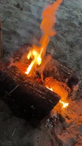 A close-up shot shows a small campfire burning on sandy ground. Three thick, dark logs are arranged in a rough triangle, with bright orange and yellow flames rising from their center. Wisps of white smoke curl upwards from the fire. Red embers and gray ash are visible at the base of the burning logs. The sandy ground is light brown. The overall lighting suggests it is dusk or night, with the fire providing the primary illumination.