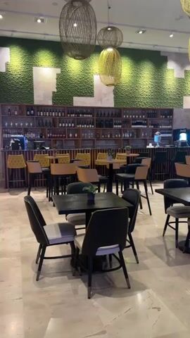 This video pans across a modern restaurant or lounge area. The space features a green textured wall with ambient lighting, a bar area with shelves stocked with bottles, and various seating arrangements. There are several dark-colored square tables with black chairs, some with light-colored seat cushions, scattered throughout the room. In the foreground, a few tables are clearly visible. Further back, more seating areas and bar stools are present. Decorative elements include large, woven pendant lights hanging from the ceiling and a small cart with decorative items in the middle of the room. The flooring is a light-colored polished stone or tile. The overall atmosphere appears to be calm and unoccupied.