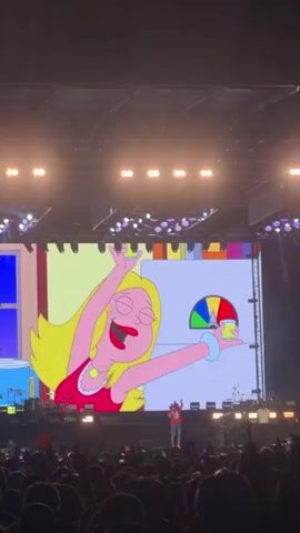 The video shows a large screen displaying animated characters from the TV show American Dad at a live concert. In the foreground, a silhouette of a performer and a crowd of people are visible. The screen displays various scenes, including Francine Smith dancing, Roger Smith interacting with her, Stan Smith looking concerned, and another animated character with a red body holding drinks. The animation is bright and colorful, with scenes depicting what appear to be domestic settings with exaggerated elements. The overall impression is that of a concert where animated visuals are being used as part of the performance.