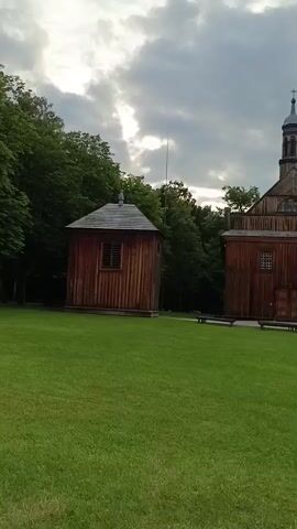 The video opens to a shot of a small wooden building with a pitched roof surrounded by a manicured green lawn and a dense line of trees in the background. The sky above is cloudy with patches of sunlight breaking through. The camera pans right, revealing a row of wooden structures behind the trees, suggesting a village or historical exhibit. The pan continues, showing the initial wooden building again, then a larger wooden structure resembling a church with a steeple. The camera then pans further right to reveal another small wooden building and then a wider shot of the grassy area with a person sitting in the distance. The camera continues to pan, showing more trees and greenery. The video appears to be a walk-through of an outdoor historical village or museum.