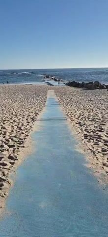 The video shows a sandy beach with a blue pathway leading towards the ocean. The sun is shining brightly, creating a glare on the water. There are a few people on the beach, some walking and others sitting under umbrellas. In the distance, there are rocks and waves crashing on the shore.