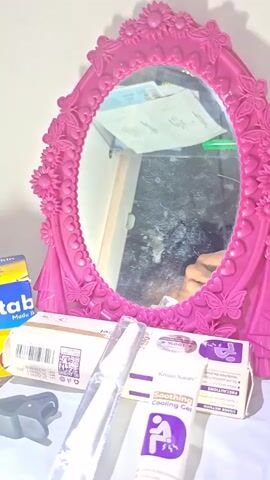 A pink ornate oval mirror with floral and butterfly accents is the central focus. To the left of the mirror are various items on a white surface, including a yellow and blue box, a black clip, and two tubes of a purple and white packaged product labeled "Soothing Cooling Gel." To the right of the mirror, a smartphone is propped up, displaying a video or image of a city street scene with buildings and cars. The mirror's reflection shows a cluttered surface with papers and a glimpse of a person's hand. The lighting is dim, casting a soft glow on the scene.