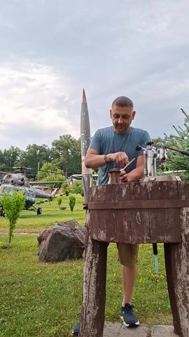 The media file shows a man standing outdoors, with a large projectile, possibly a missile, visible behind him. He is wearing a blue t-shirt and khaki shorts. The man is looking down at something he is holding in his hands, which appears to be a small wooden object. He has a slight smile on his face.

The setting is a grassy area with trees in the background. There are other objects that look like military equipment, including what appears to be a helicopter and another large projectile, partially visible in the distance. A large rock is also present in the foreground. The sky is overcast, suggesting it is daytime, possibly late morning or afternoon.

The man's posture and facial expression suggest he is engaged in a task or perhaps showing off an object. The presence of military equipment in an outdoor setting might indicate a museum, a historical site, or a memorial park. The overall scene has a casual, outdoor feel. There is no visible text in the media file.