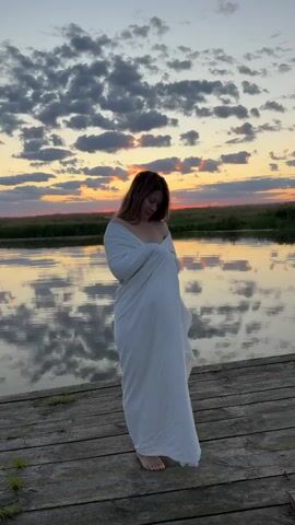 A young woman stands on a wooden dock, wrapped in a white sheet. She is facing a body of water that reflects the sky, which is filled with dramatic clouds illuminated by a sunset. The woman is looking away from the camera towards the sunset. The scene is tranquil and serene.
